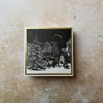 A square framed art print featuring a black and white winter village scene with a Christmas tree and stars, displayed on a textured beige wall.