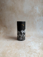 A small cylindrical container or tin with a black lid and a marbled black-and-white body, standing upright on a textured beige surface against a matching beige backdrop.