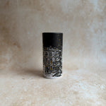 A small cylindrical candle holder or tumbler with a black top and a detailed black-and-white winter house illustration on the lower half, set against a textured beige background.