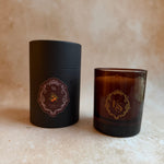 An amber glass candle jar next to its matte black cylindrical packaging box, both featuring a gold and pink circular monogram logo with the letters 'KS'.