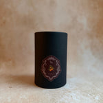 A matte black cylindrical tin container with a decorative circular label, set against a textured beige stone-like background.