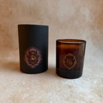 An amber glass candle jar standing next to its matte black cylindrical packaging container, both featuring a gold circular monogram logo with the letters 'KS'.