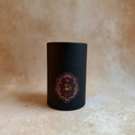 A matte black cylindrical tin container with an ornate circular label, set against a textured beige stone background.