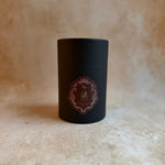 A matte black cylindrical tin container with a circular ornate label in burgundy and gold, standing on a textured beige stone surface against a matching beige background.