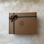 A square brown kraft paper gift box tied with a dark green ribbon and decorated with a dried orange slice tag, placed on a textured beige surface.