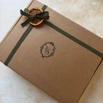 A square kraft paper gift box tied with an olive green ribbon and decorated with a dried orange slice on a light beige surface.